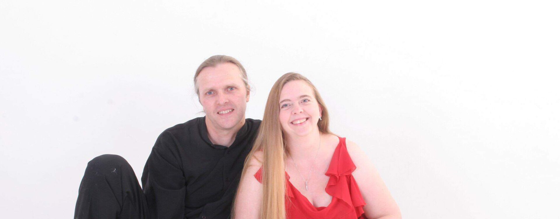 A man dressed in all black and a woman in a red dress with long blonde hair.  They're both smiling and facng the camera