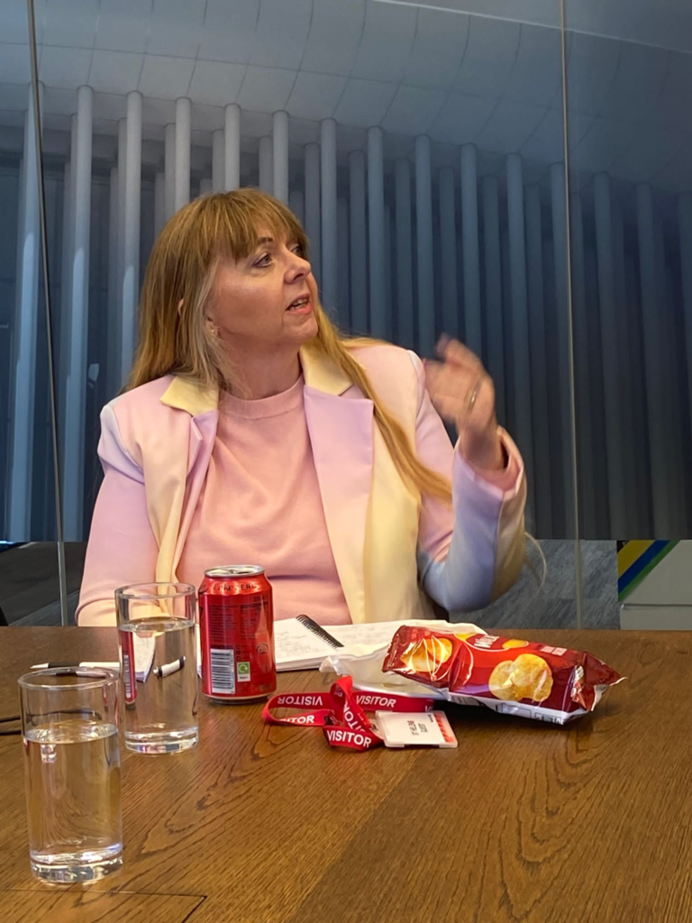 Image is of Leslie speaking at the ADHD and women event.  Leslie is seated at a conference table and is wearing a pastel rainbow coloured taliored jacket and a light pink jersey. 