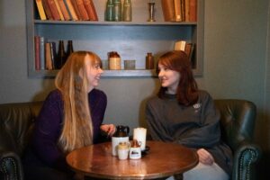 A photo of a woman and her daughter looking at each other. They are seated at a table with books behind them. They are laughing companionably with each other