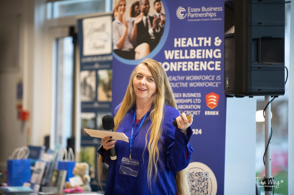 Picture of Leslie with a microphone in hand, wearing a blue sating top.  She is standing in front of an advertising banner for Essex Business Partnership and the wellbeing conference where she spoke about ADHD in the workplace