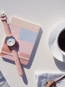 Image of a pink patchwork notebook with a pink watch lying on top of it. On the right is a cut off cup of black coffee in a white china cup and saucer